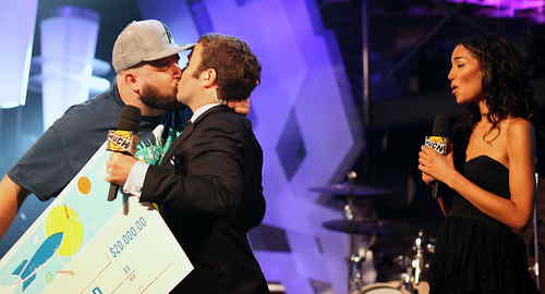 Pink Eye (in trucker hat) plants a kiss on Grant Lawrence (in suit) while a hostess (in evening gown) looks on, shocked